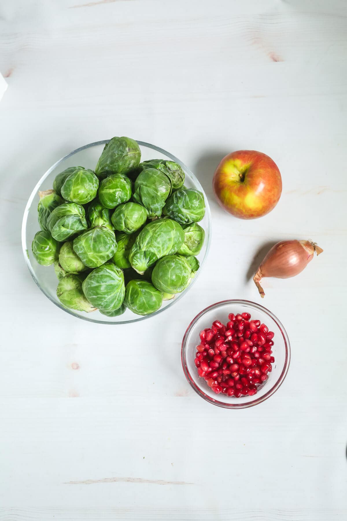 A glass bowl displays a Brussels Sprouts Shaved Salad, elegantly accompanied by a whole apple, a shallot, and a small bowl of pomegranate seeds on a pristine white surface.