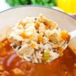 A ladle holding a portion over a pot reveals tender chicken, rice, and vibrant vegetables. Fresh herbs and a yellow bell pepper blur softly in the background, enhancing this flavorful Cajun delight with aromatic charm.