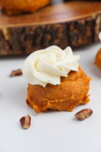 A pumpkin-flavored dessert made with Cake Mix Pumpkin Spice Cookies, topped with a swirl of white frosting and placed on a white surface with pecan pieces nearby.