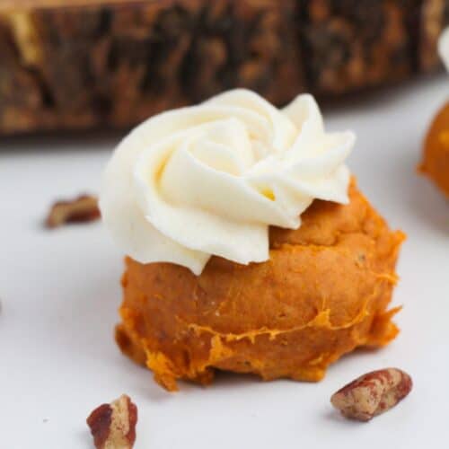 A pumpkin-flavored dessert made with Cake Mix Pumpkin Spice Cookies, topped with a swirl of white frosting and placed on a white surface with pecan pieces nearby.