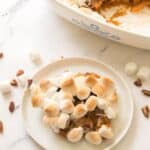 A plate with a serving of candied sweet potatoes with canned sweet potatoes casserole topped with toasted marshmallows, next to a baking dish, scattered pecans, and marshmallows on a white surface.