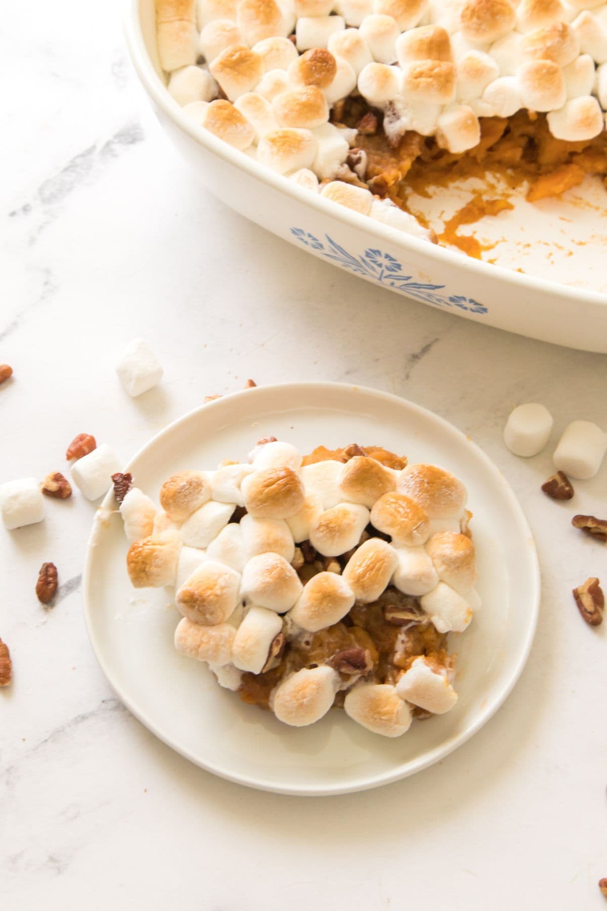 A plate with a serving of candied sweet potatoes with canned sweet potatoes casserole topped with toasted marshmallows, next to a baking dish, scattered pecans, and marshmallows on a white surface.