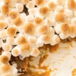 A casserole dish filled w with canned sweet potatoes, topped with toasted marshmallows, with a portion already served.