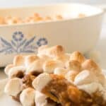 A serving of candied sweet potatoes with canned sweet potatoes, topped with toasted marshmallows on a plate, with a casserole dish in the background.