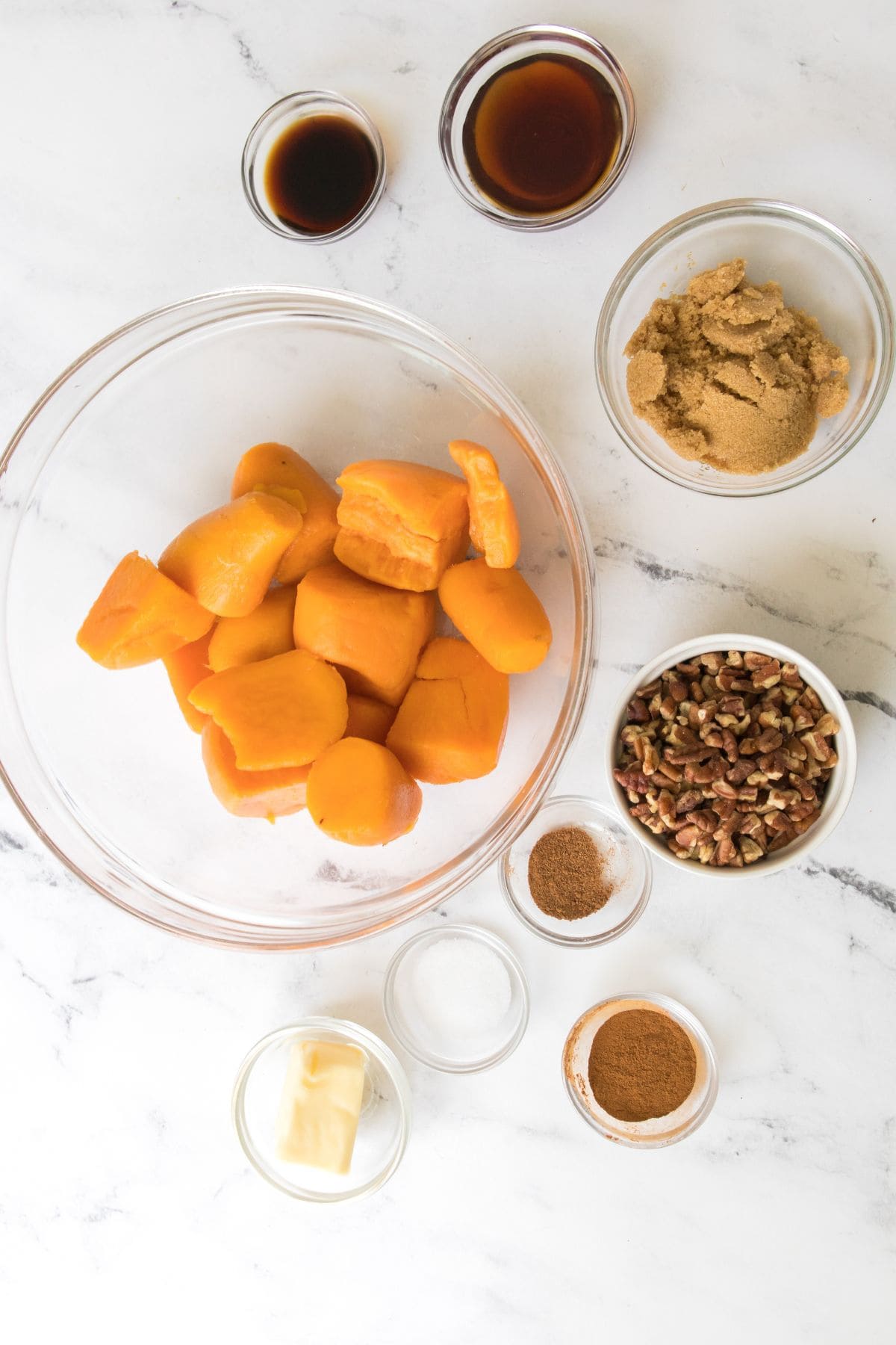 A glass bowl surrounded by small bowls of brown sugar, pecans, vanilla extract, cinnamon, nutmeg, butter, and syrup on a white marble surface.