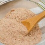 A glass bowl filled with a light brown, finely ground carnitas seasoning, with a wooden spoon partially submerged in the mixture.