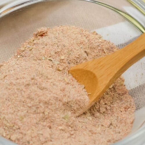 A glass bowl filled with a light brown, finely ground carnitas seasoning, with a wooden spoon partially submerged in the mixture.