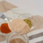 A spoonful of light brown spice held above a jar, with small bowls of green and red powders in the background on a striped cloth.