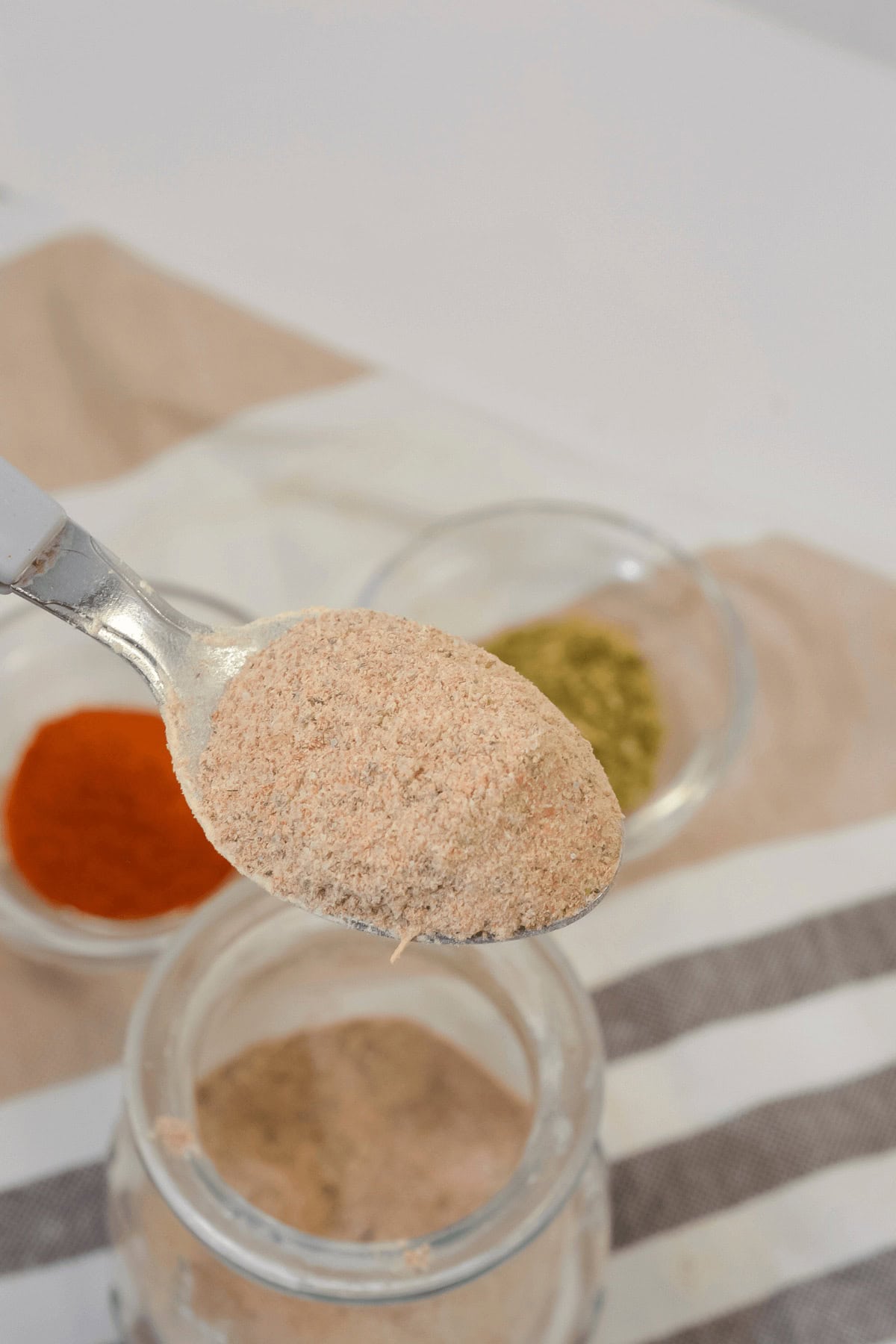 A spoonful of light brown spice held above a jar, with small bowls of green and red powders in the background on a striped cloth.