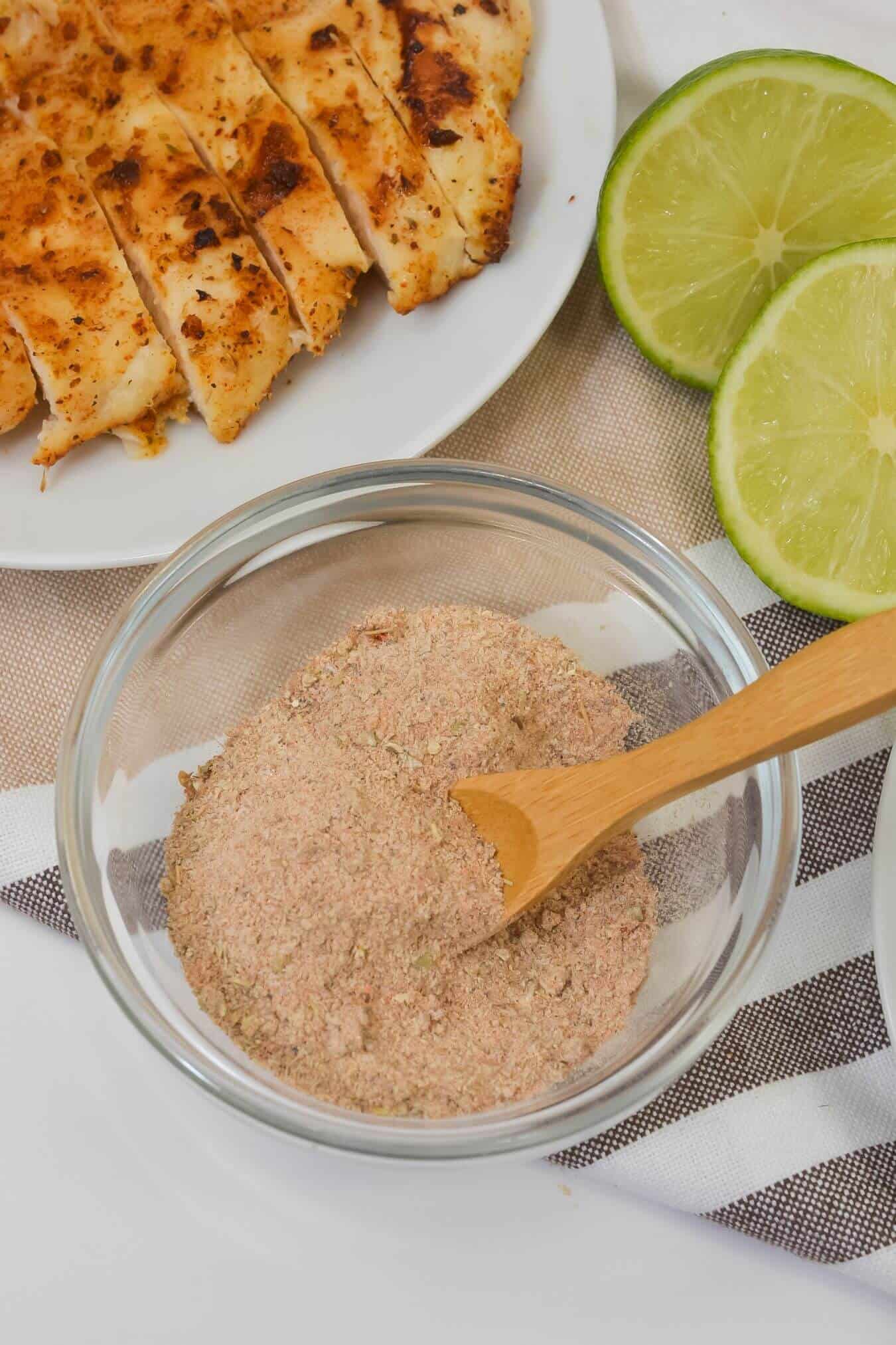 A glass bowl of mix with a wooden spoon sits next to sliced cooked chicken breast and halved limes on a table.