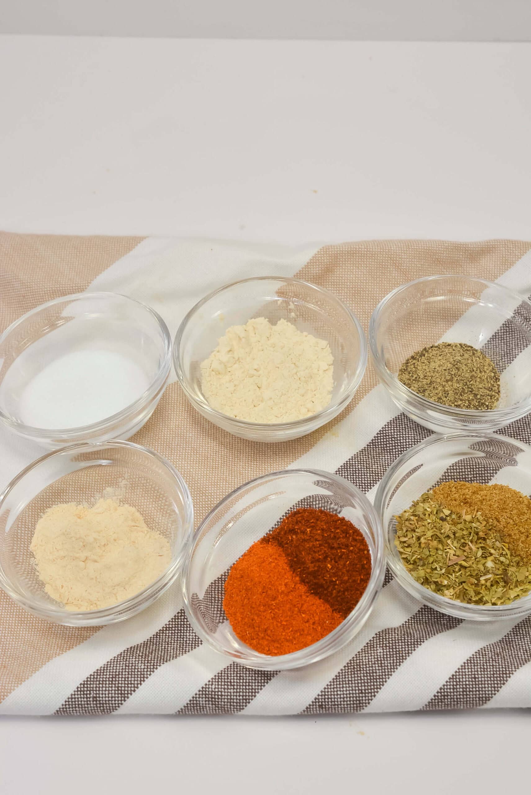 Six small glass bowls with different spices and seasonings, including salt, powders, herbs,are arranged on a striped cloth.