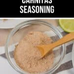 A glass bowl of flavorful carnitas seasoning with a wooden spoon rests on a striped cloth beside a fresh lime slice.