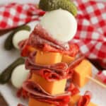 A skewer featuring a stack of deli meats, cheese cubes, a mozzarella ball, and a small pickle on top sits proudly against a checkered cloth background, embodying the essence of Charcuterie Skewers.