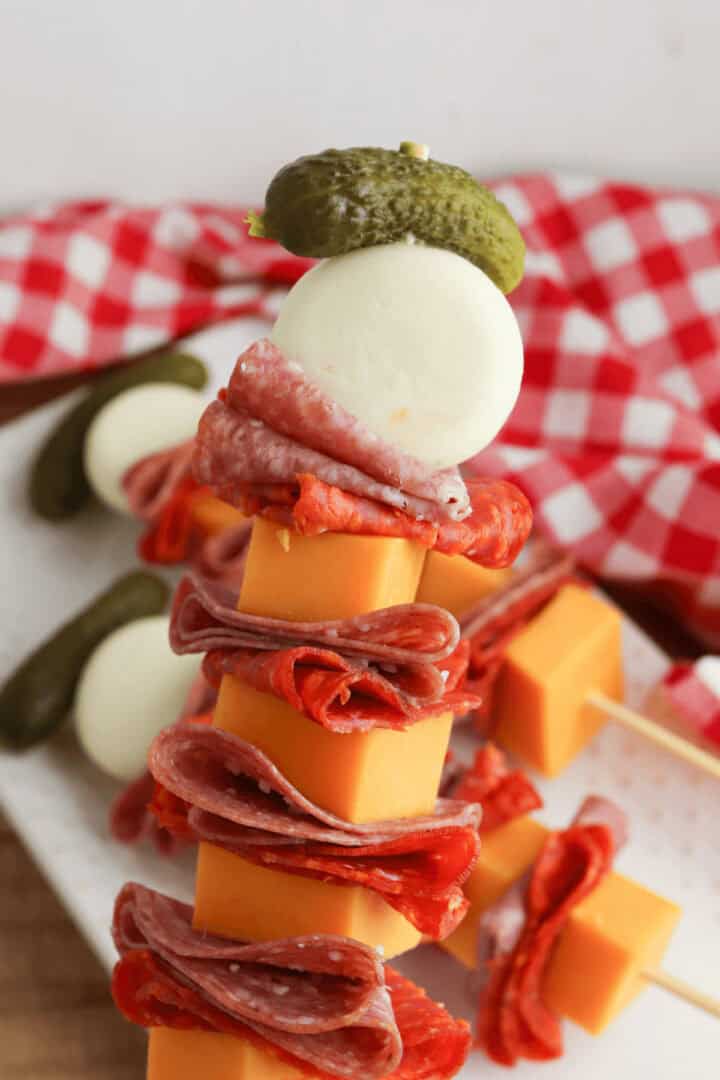 A skewer featuring a stack of deli meats, cheese cubes, a mozzarella ball, and a small pickle on top sits proudly against a checkered cloth background, embodying the essence of Charcuterie Skewers.