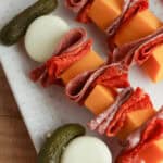 An arrangement of charcuterie skewers with cubes of cheese, slices of salami, pepperoni, boiled eggs, and pickles on a white tray.