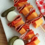 Charcuterie skewers with cheese cubes, folded slices of salami, pepperoni, a mozzarella ball, and a pickle on a white plate next to a red checkered cloth.