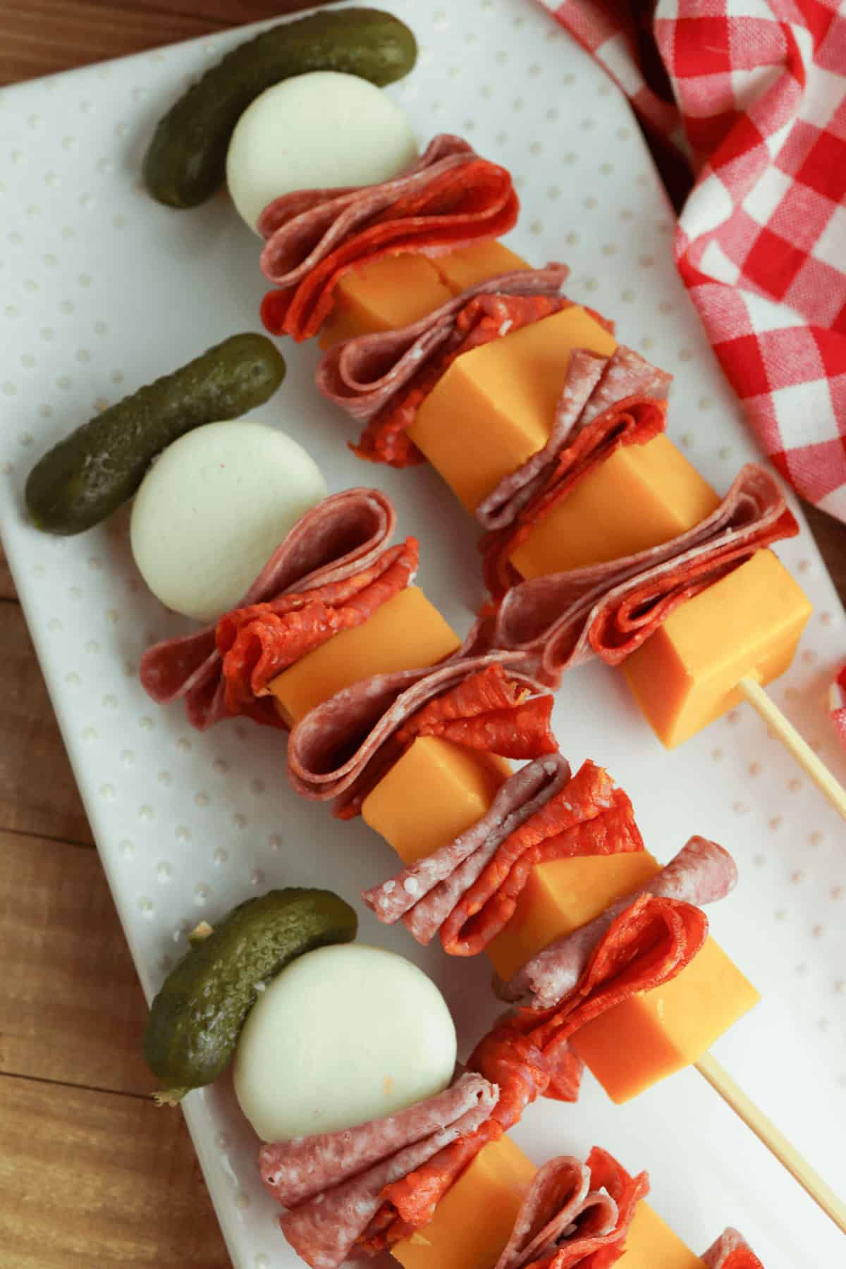 Charcuterie skewers with cheese cubes, folded slices of salami, pepperoni, a mozzarella ball, and a pickle on a white plate next to a red checkered cloth.