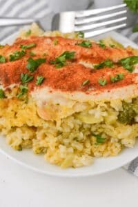 A serving of Cheesy Chicken Broccoli and Rice Casserole topped with paprika and chopped parsley on a white plate, with a fork in the background.