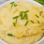 A white bowl filled with cheesy mashed potatoes, garnished with chopped chives, placed on a white surface.