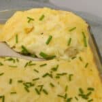 A serving of Cheesy Mashed Potatoes casserole with chopped chives on top is lifted from a baking dish.