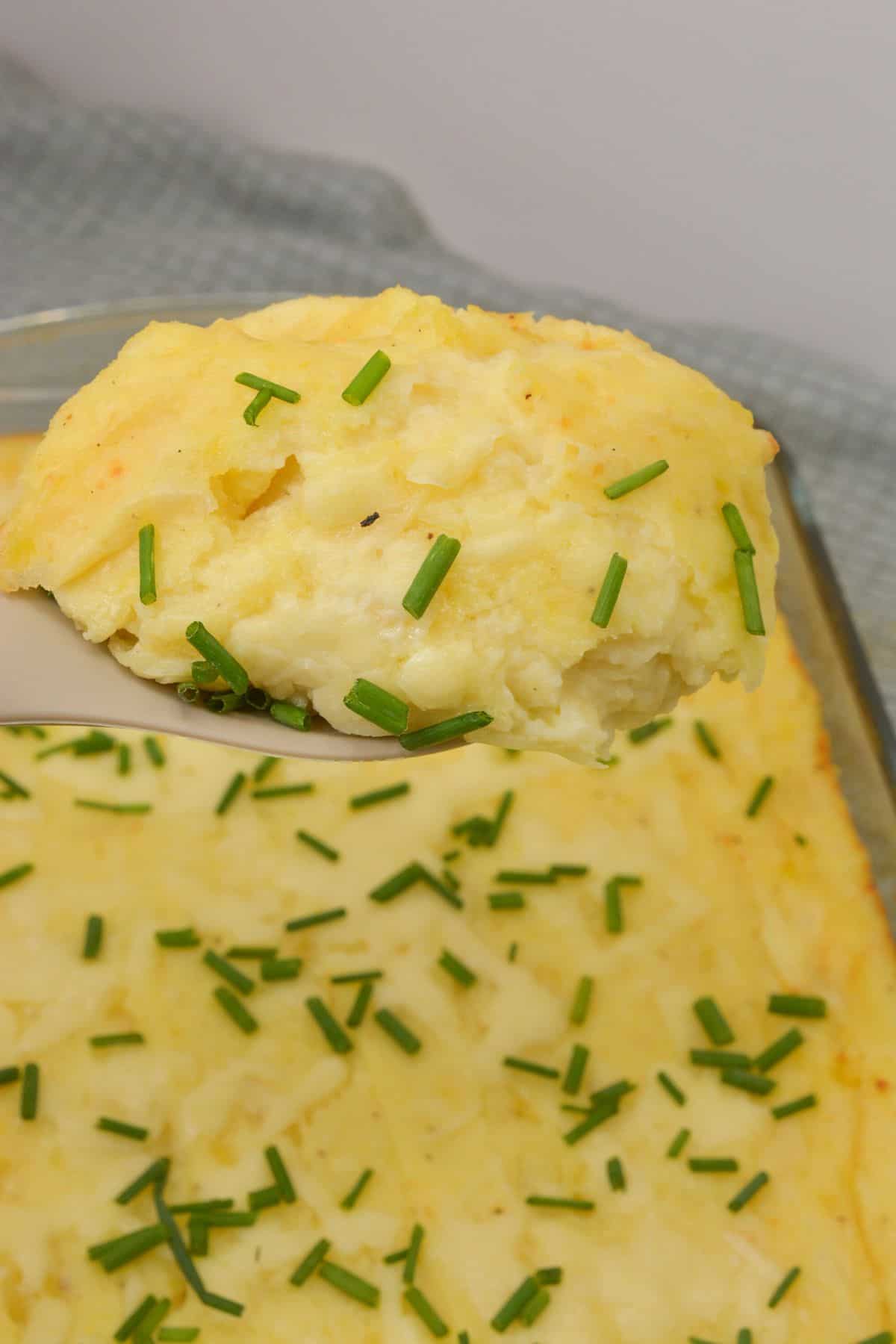 A serving of Cheesy Mashed Potatoes casserole with chopped chives on top is lifted from a baking dish.