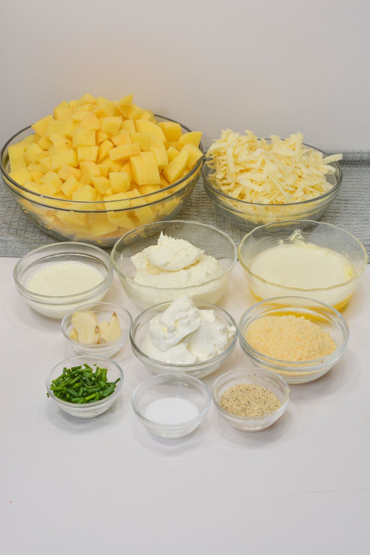 Assorted glass bowls filled with diced potatoes, shredded cheese, sour cream, cream cheese, butter, garlic, seasonings, and chopped chivesare arranged on a white surface.