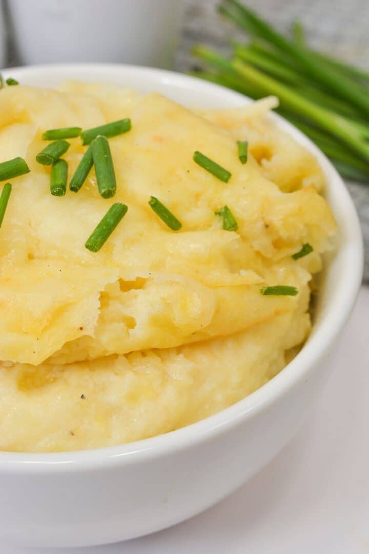 A white bowl filled with cheesy mashed potatoes, topped with chopped chives.