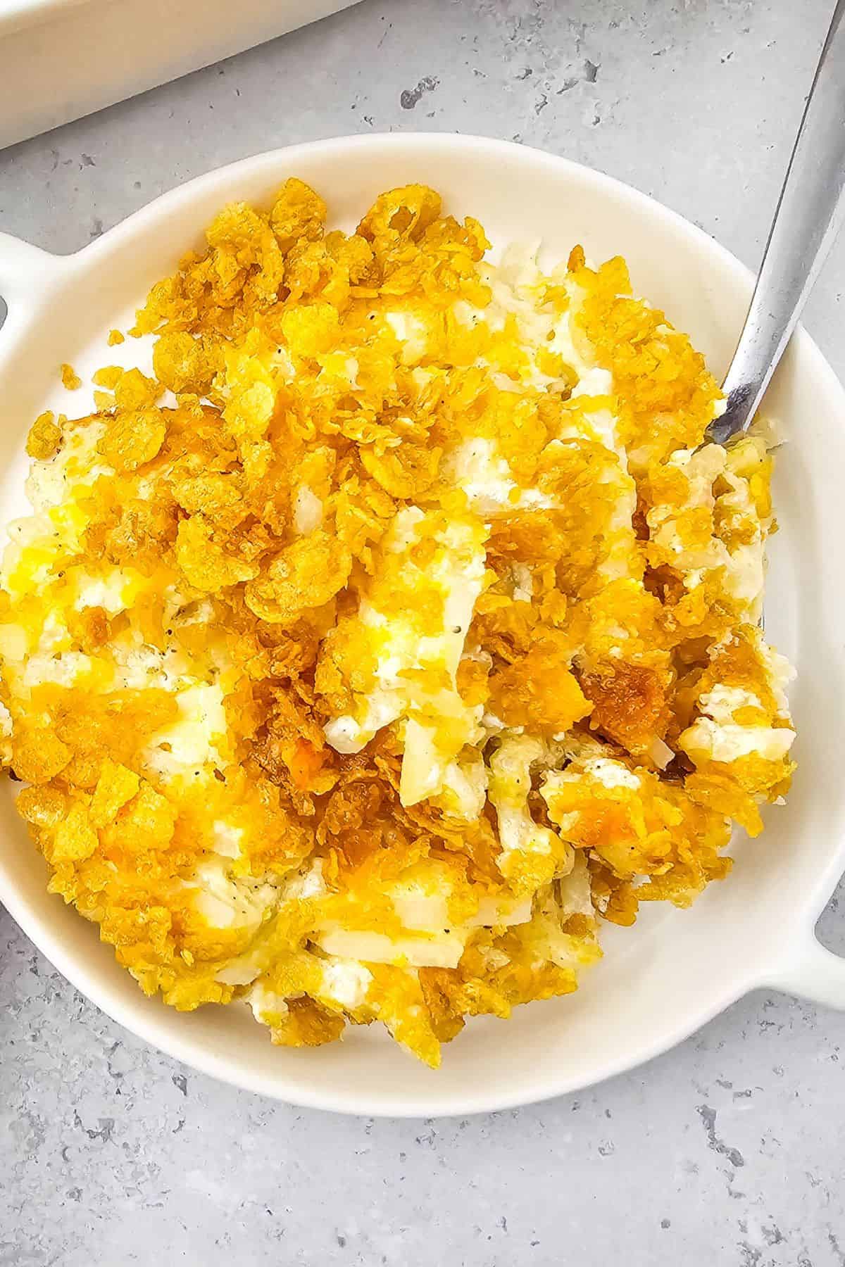 A white dish filled with Cheesy Party Potatoes casserole, topped with a crispy golden cornflake crust, with a metal spoon resting on the side.