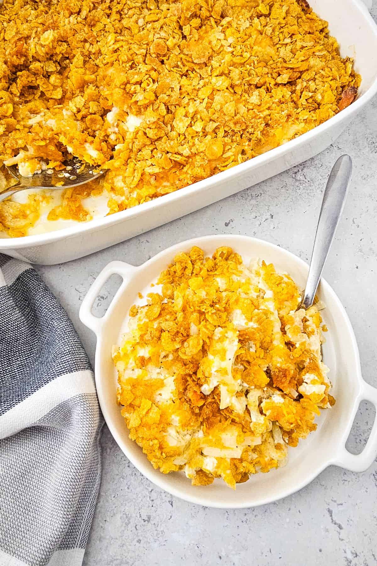 A serving of Cheesy Party Potatoes casserole with a crispy cornflake topping in a white dish, next to a casserole dish and a striped cloth napkin.