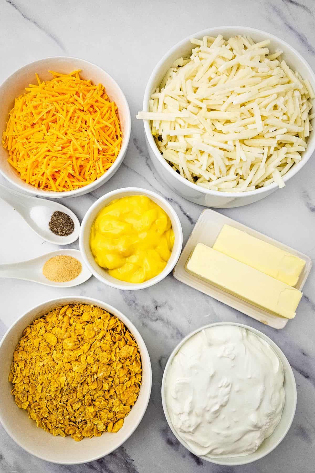 Bowls containing ingredients for shredded cheddar cheese, hash browns, crushed corn flakes, sour cream, condensed soup, butter sticks, plus small spoons of pepper and garlic powder—sit on a marble counter.