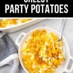 A white dish filled with Cheesy Party Potatoes, topped with a crispy golden layer, sits on a light surface with a spoon inside and a striped towel beside it.