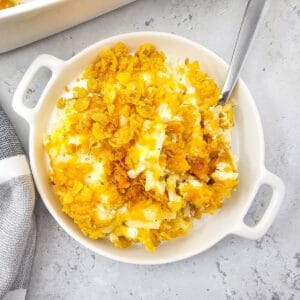 A round white dish filled with creamy Cheesy Party Potatoes casserole, topped with golden, crispy cornflakes, with a spoon resting inside the dish.