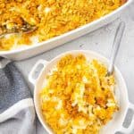 A serving of Cheesy Party Potatoes—classic hashbrown casserole with a crispy cornflake topping—in a bowl beside a larger baking dish, both with metal spoons on a light countertop.