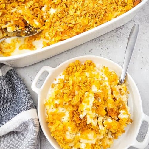 A serving of Cheesy Party Potatoes—classic hashbrown casserole with a crispy cornflake topping—in a bowl beside a larger baking dish, both with metal spoons on a light countertop.