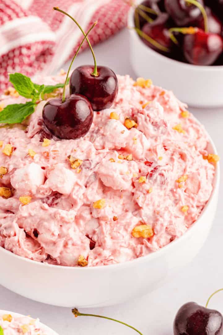 A white bowl filled with Cherry Fluff Salad, topped with two cherries and mint, sits in front of a bowl of fresh cherries and a red cloth in the background.