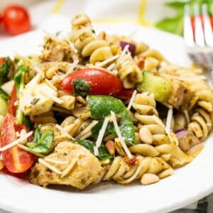 A plate of Chicken and Pesto Pasta Salad with rotini, grilled chicken, cherry tomatoes, spinach, zucchini, pine nuts, and shredded cheese.