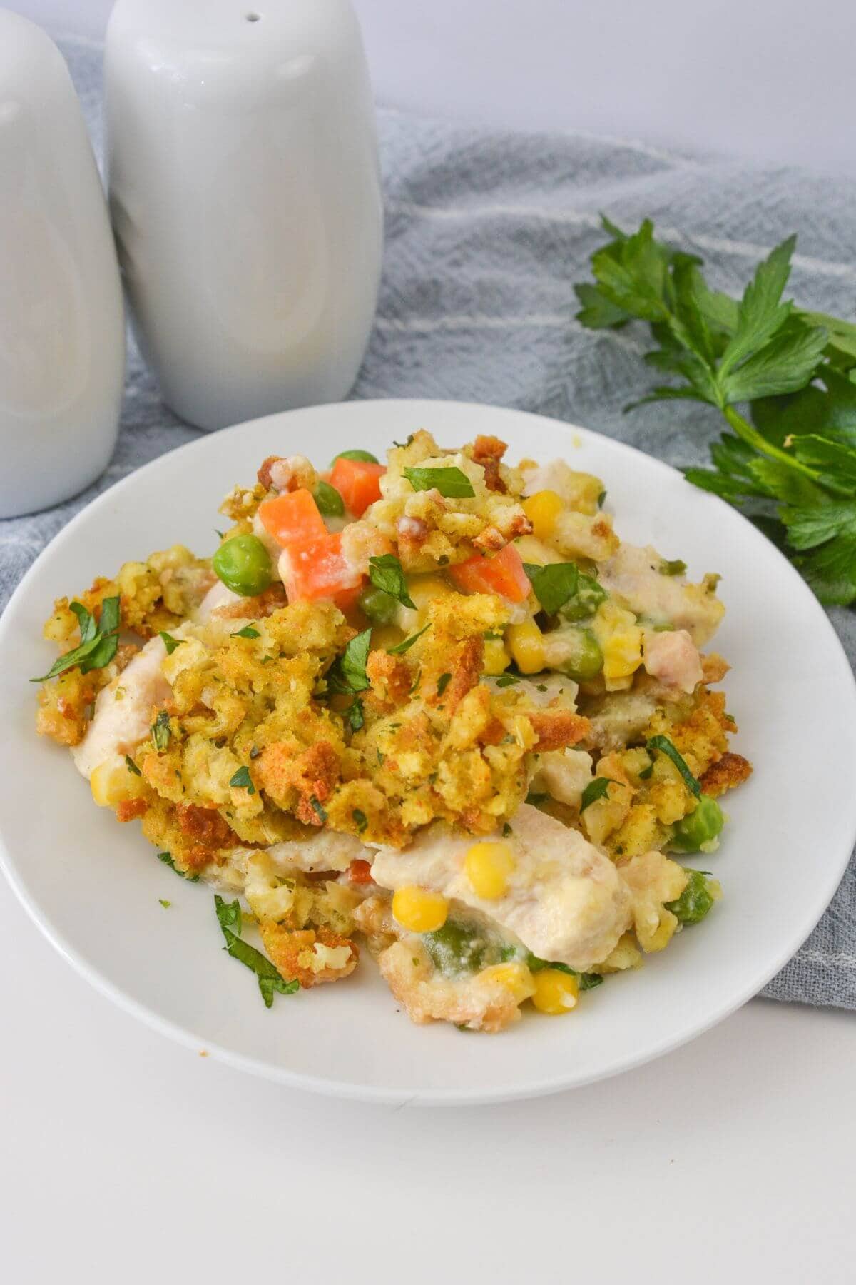 A delightful plate of chicken and stuffing casserole accompanied by mixed vegetables. Peas, carrots, and corn are garnished with parsley, all beautifully presented on a white dish next to two sleek white salt and pepper shakers.