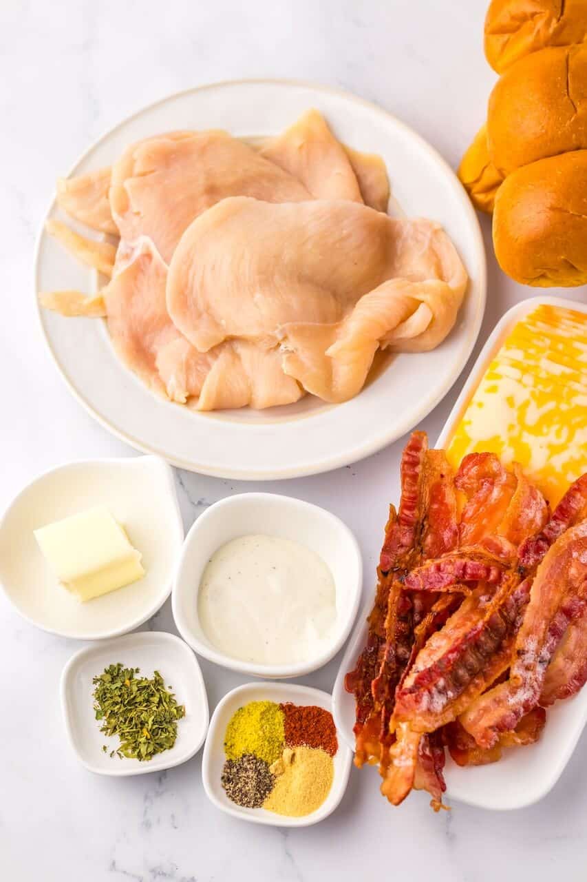 Image of ingredients including raw chicken breasts on a plate, crispy bacon strips, butter, various spices, sauce, and bread rolls on a marble countertop, perfect for making Chicken Bacon Ranch Sliders.