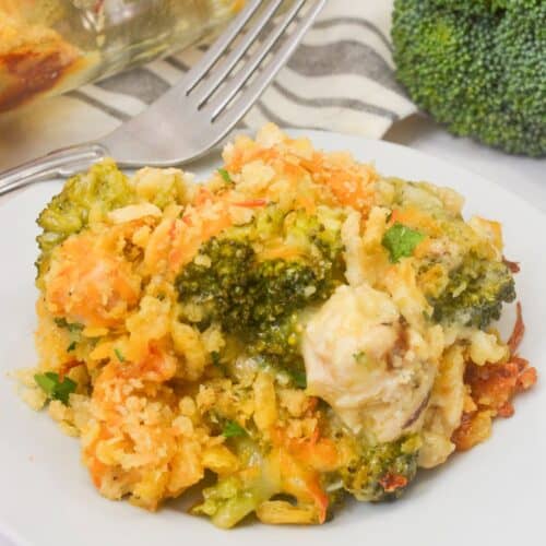 A serving of Chicken Broccoli Divan casserole on a white plate with a fork, next to fresh broccoli and a baking dish.