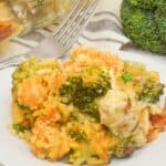 A serving of broccoli and cheese casserole, reminiscent of Chicken Broccoli Divan, sits on a white plate with a fork, next to a head of fresh broccoli and a glass baking dish in the background.