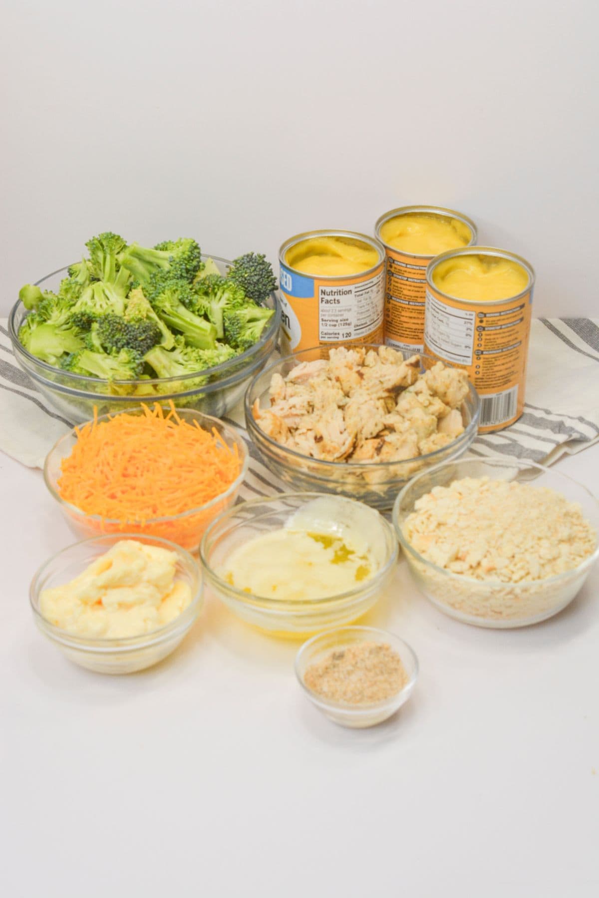 Ingredients are arranged on a white surface, including broccoli, canned soup, shredded cheese, cooked chicken, crushed crackers, mayonnaise, and seasonings in glass bowls and cans.