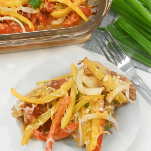 A plate with a serving of Chicken Fajita Casserole covered in melted cheese, sliced yellow and red bell peppers, and onions. A baking dish with the same delicious casserole and a bunch of green onions are in the background.