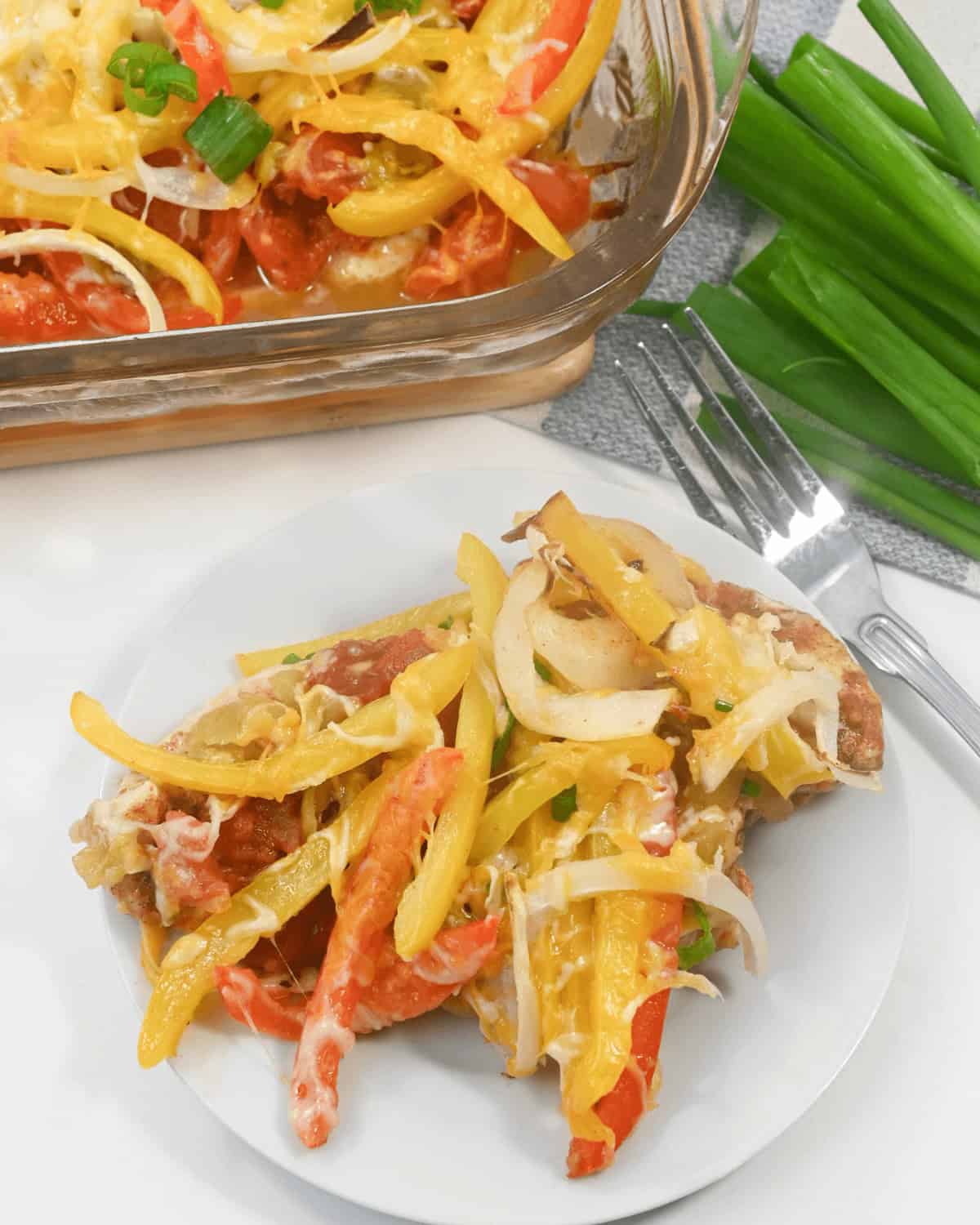A plate with a serving of Chicken Fajita Casserole covered in melted cheese, sliced yellow and red bell peppers, and onions. A baking dish with the same delicious casserole and a bunch of green onions are in the background.