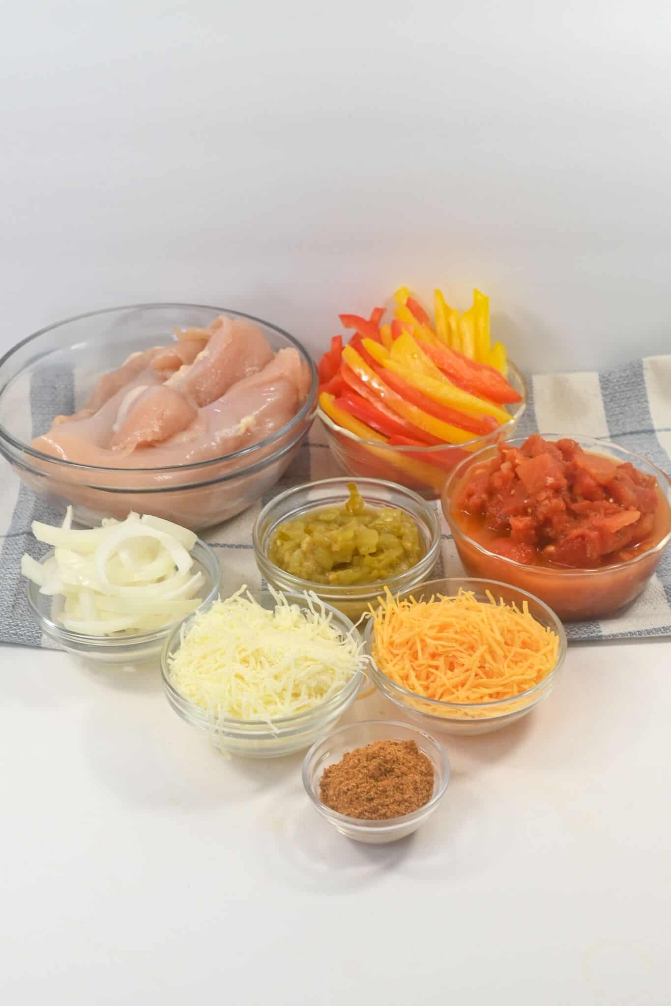 Raw chicken strips, sliced bell peppers, diced tomatoes, shredded white and yellow cheese, sliced onions, chopped green peppers, and a small bowl of seasoning—all essentials for a arrayed in glass bowls on a white surface.