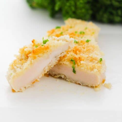 A breast, breaded and baked to perfection then sliced in half to reveal its juicy inside, with a bunch of leafy green parsley in the background.