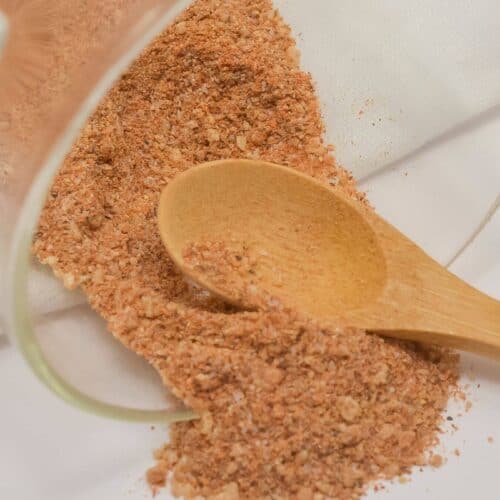 A wooden spoon rests on a white surface beside a pile of reddish-brown Chicken Wing Seasoning Rub, spilled from a glass container.