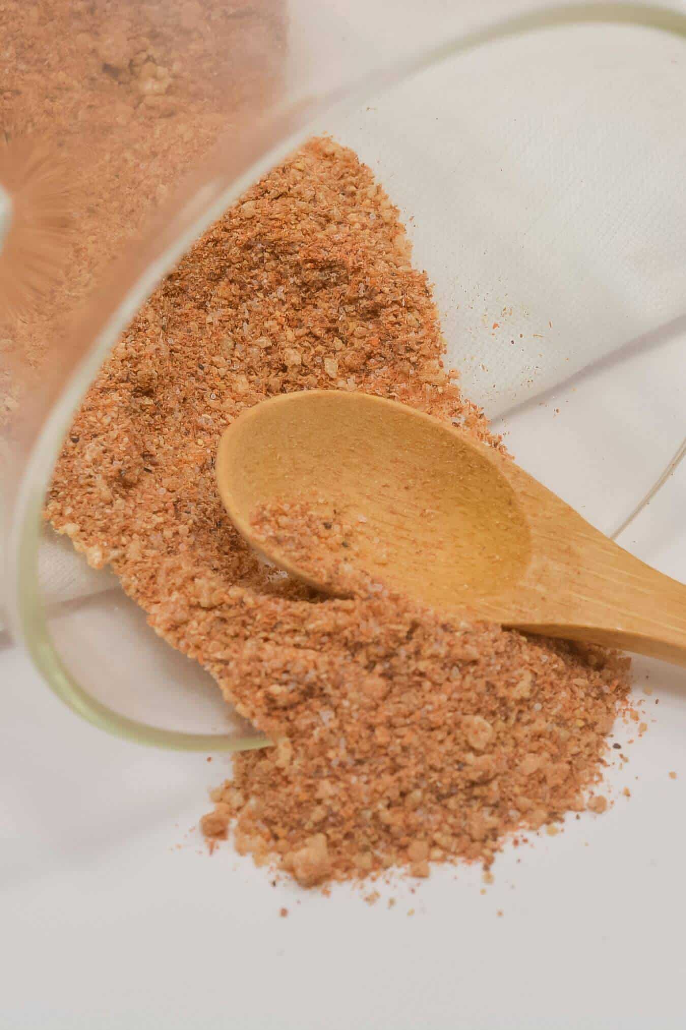 A wooden spoon rests on a white surface beside a pile of reddish-brown Chicken Wing Seasoning Rub, spilled from a glass container.
