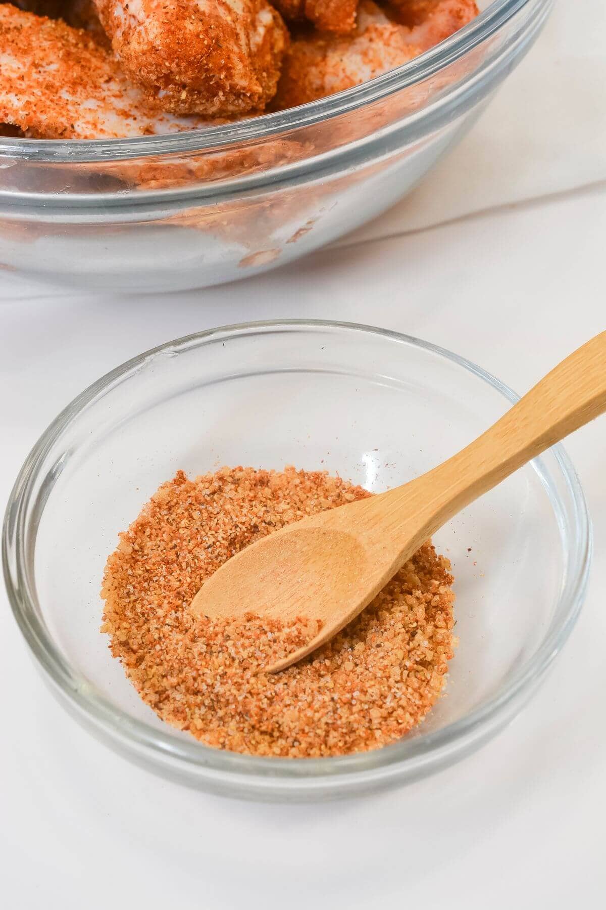 A small glass bowl containing Chicken Wing Seasoning Rub with a wooden spoon, placed in front of a larger bowl with seasoned food.