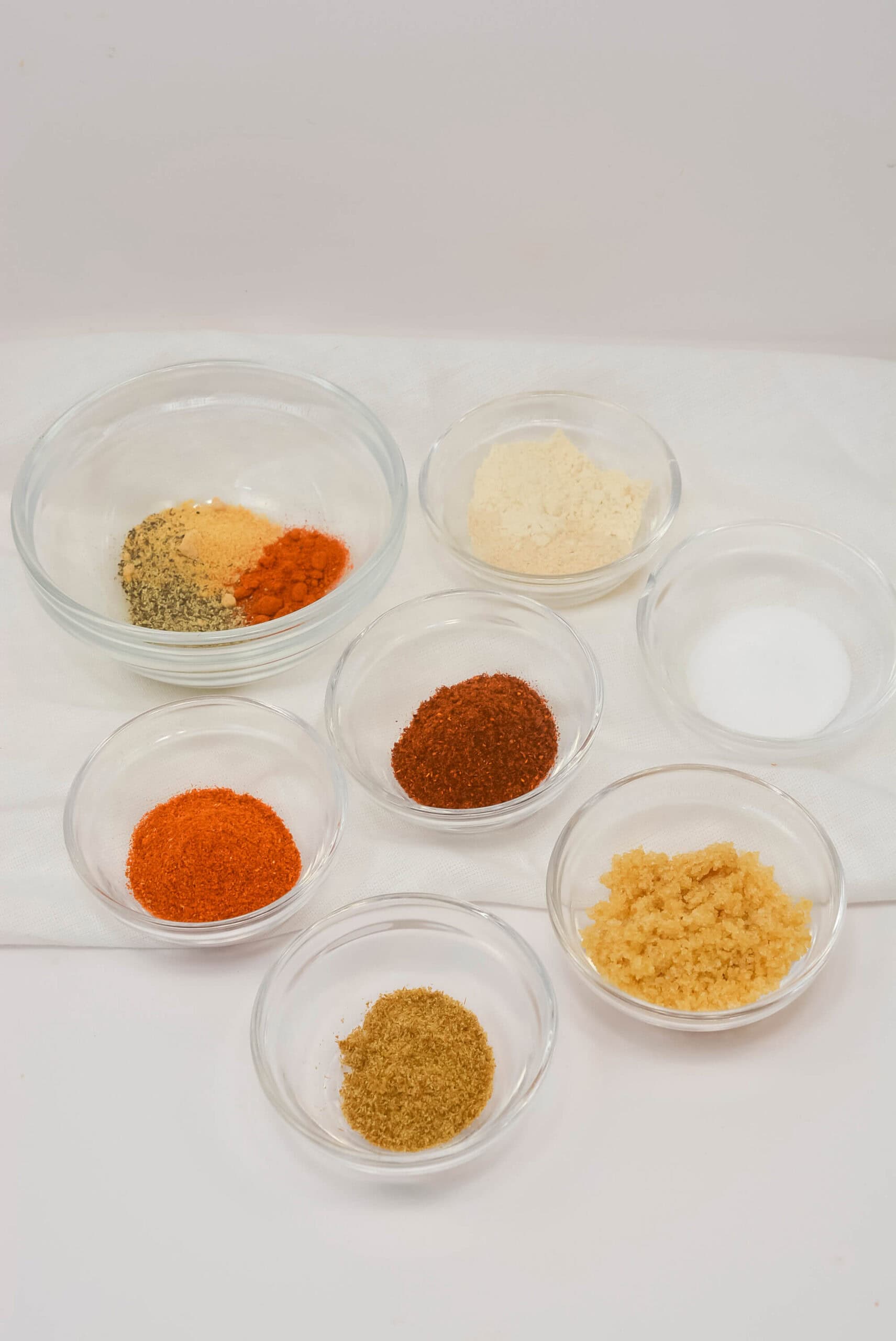 Eight small glass bowls filled with various ground spices and powders, perfect for creating your own chicken wing seasoning rub, are arranged on a white surface.