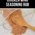 A jar of Chicken Wing Seasoning Rub is tipped over, spilling the flavorful spice blend onto a white surface with a wooden spoon resting in the vibrant pile.